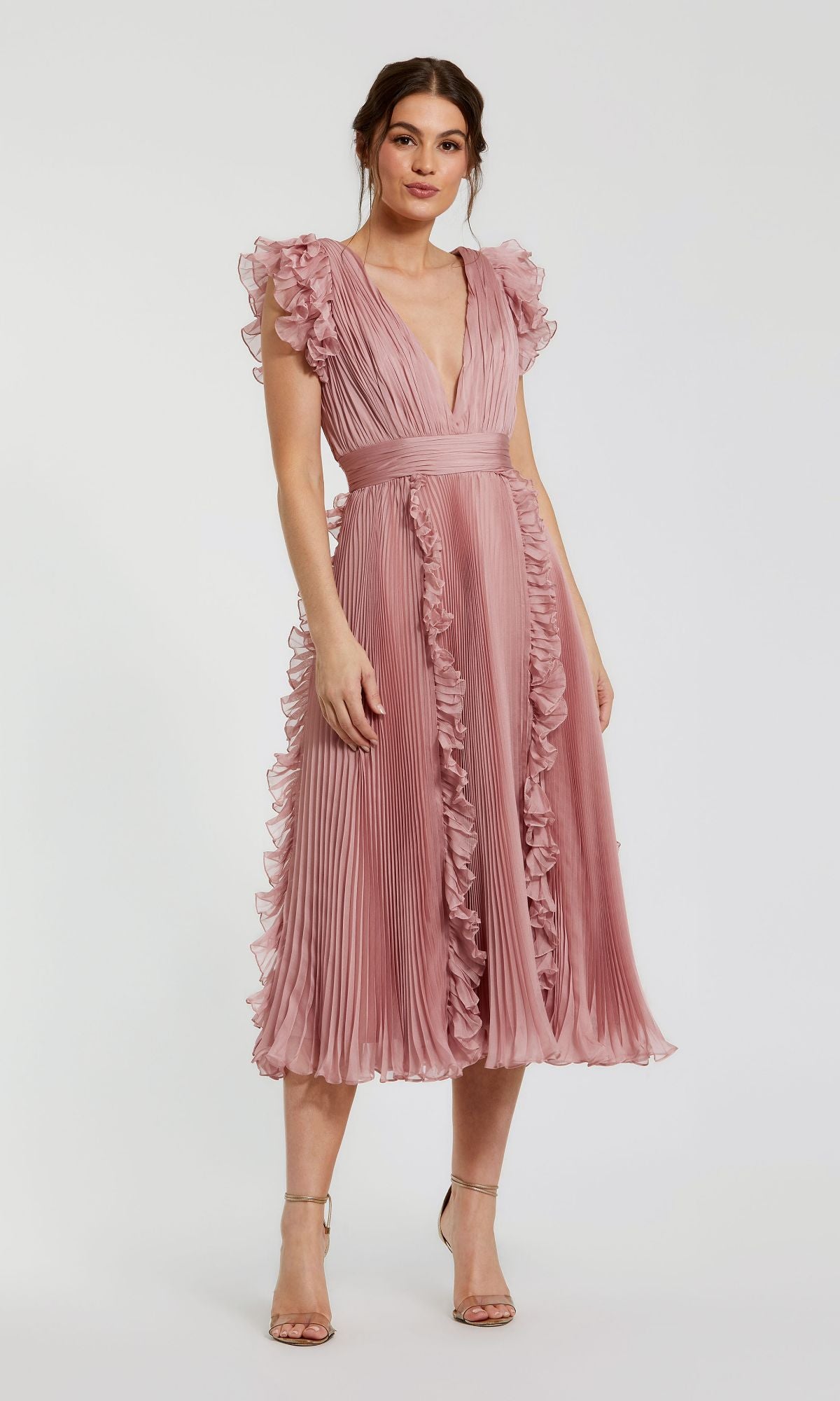 A woman wears the Short Wedding Guest Dress 49675 by Mac Duggalโa rose pink chiffon, pleated knee-length dress with ruffle cap sleeves and a V-necklineโpaired with nude heels, standing against a plain background.